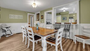 A dining area with a wooden table and white chairs, a high chair, and a vase with flowers.