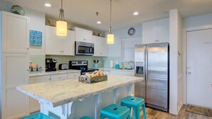 A modern kitchen with white cabinets, a marble island, teal bar stools, a stainless steel refrigerator, and a microwave.
