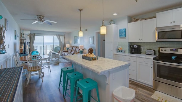 A modern kitchen with white cabinets, a large island with bar stools, a ceiling fan, and a view of the ocean through large windows.