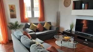 A living room with a grey sofa, a glass coffee table, a flat-screen TV, and orange curtains.