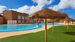A swimming pool with a thatched roof structure, a building with multiple windows, and a grassy area. - ATTRACTIVE FRONT LINE detached villa with SUPERB Jacuzzi & Tiki Rooftop Bar (Corralejo)