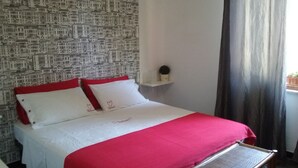 Une chambre d’hôtel avec un papier peint à motifs, un lit avec une literie rouge et blanche, une petite étagère ornée d’un objet décoratif et une fenêtre avec des rideaux.
