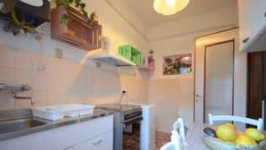 Une cuisine avec des meubles blancs, un évier en acier inoxydable, un four encastré et un coin repas comprenant une chaise blanche et un panier de fruits.