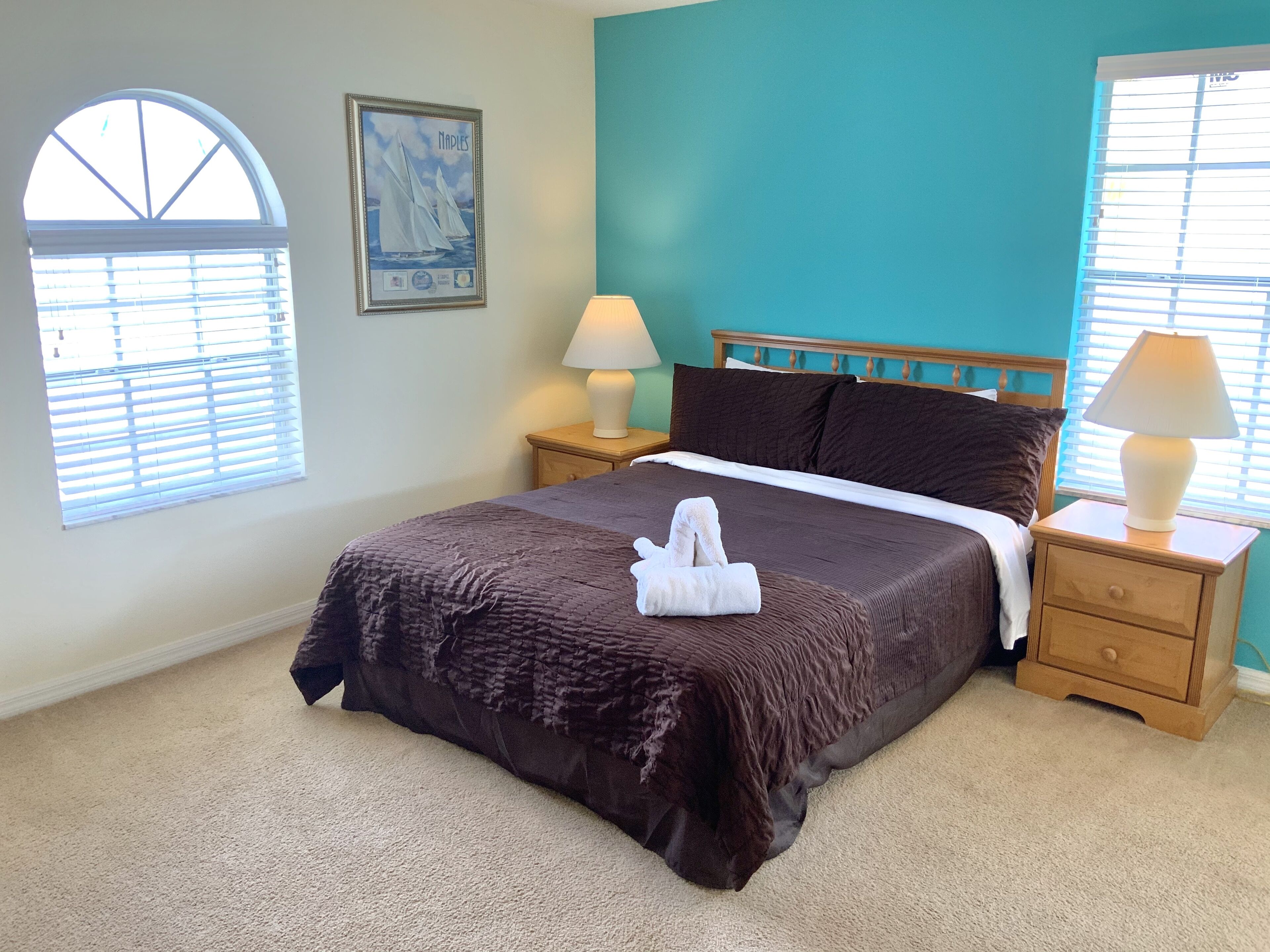 A neatly made bed with a brown bedspread and a white towel, two bedside lamps, a wooden nightstand, a framed picture on the wall, and a window with blinds.