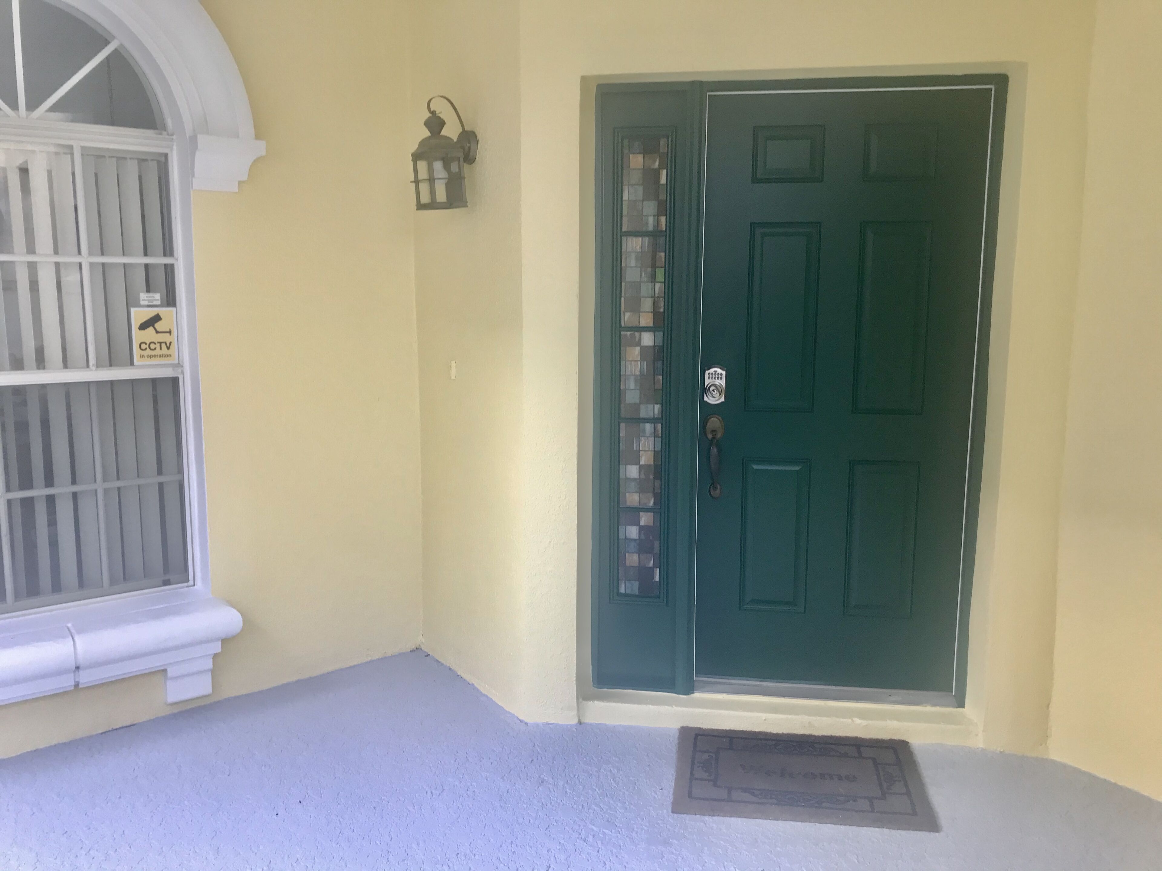A green door with a doormat, a CCTV sign, and a wall-mounted lantern.