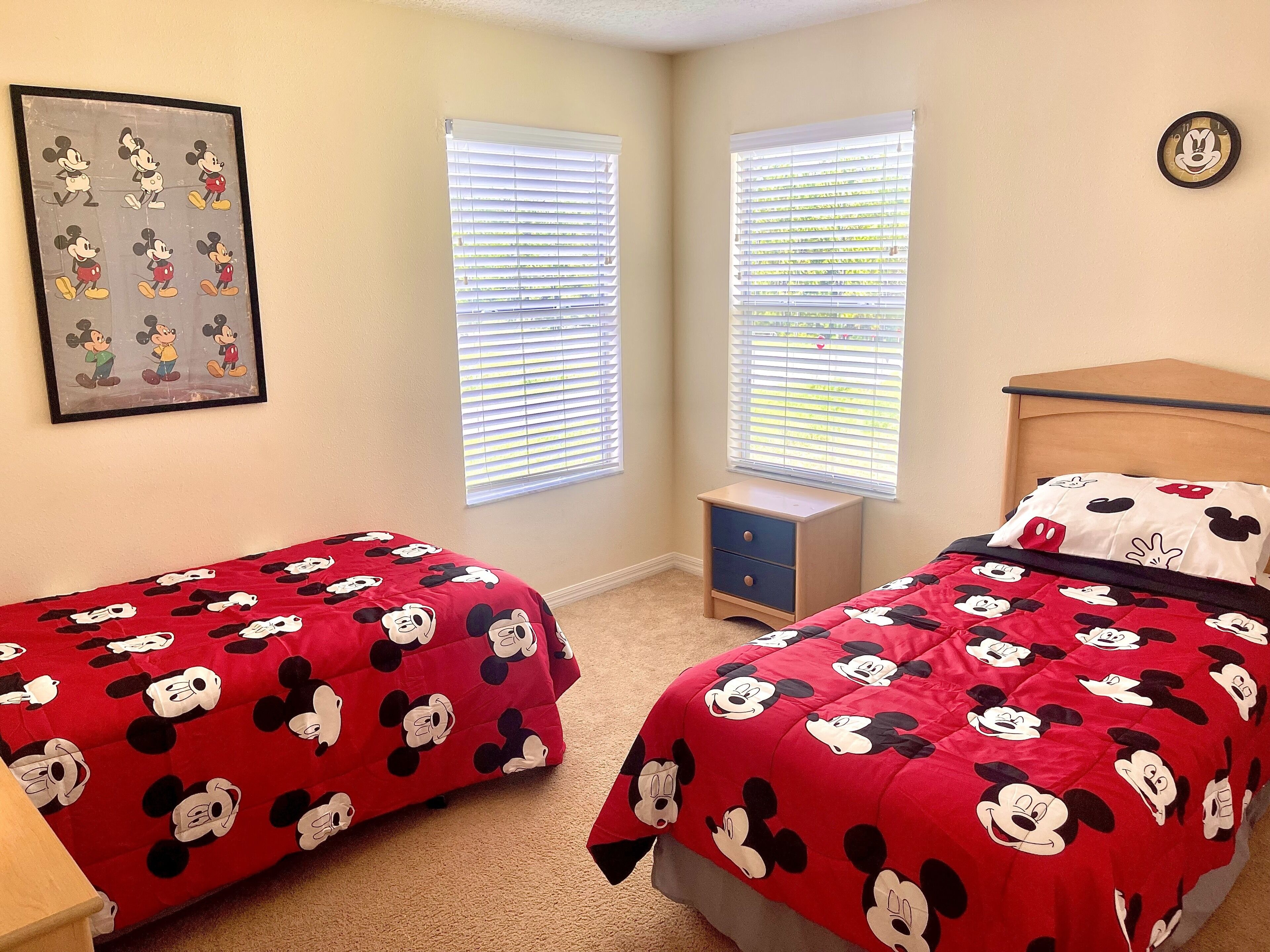 Une chambre avec deux lits, chacun recouvert d’une couverture aux motifs de Mickey Mouse. On y trouve une petite table de chevet avec une lampe, un tableau encadré au mur et une horloge au mur.