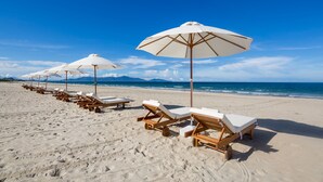 A beach with rows of wooden lounge chairs and white umbrellas under a clear blue sky. - Grandvrio Ocean Resort Danang (Da Nang)