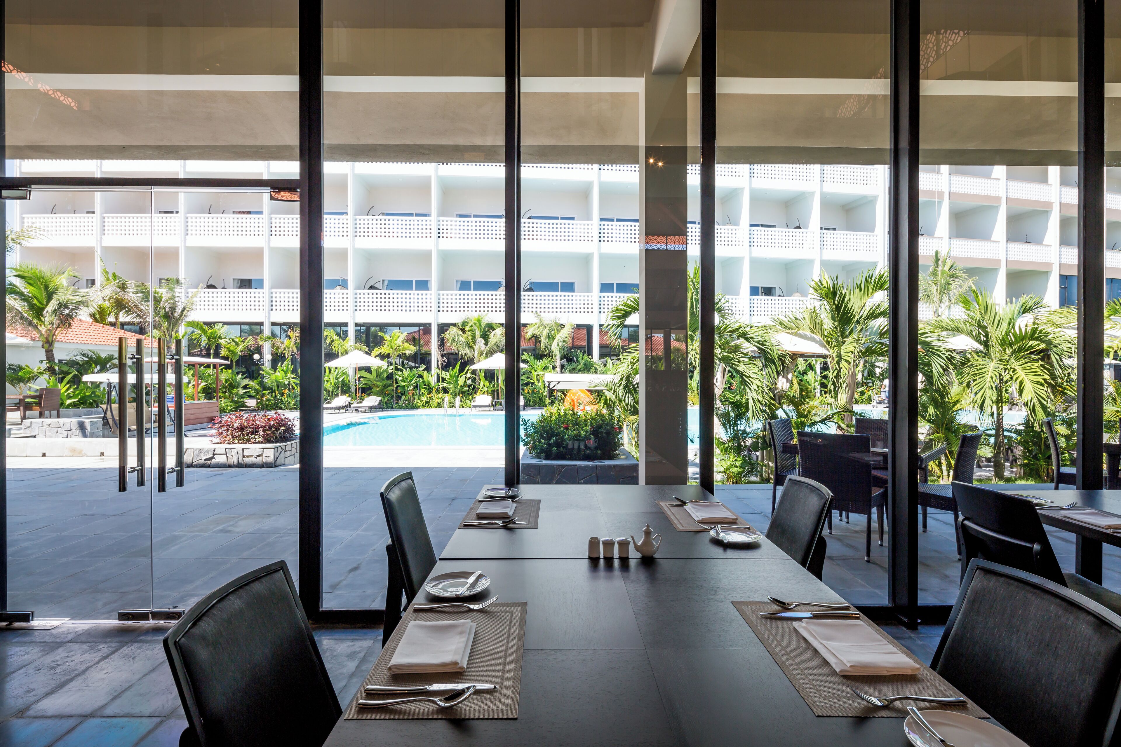 A dining area with tables set for a meal, overlooking a pool and a landscaped garden with palm trees.