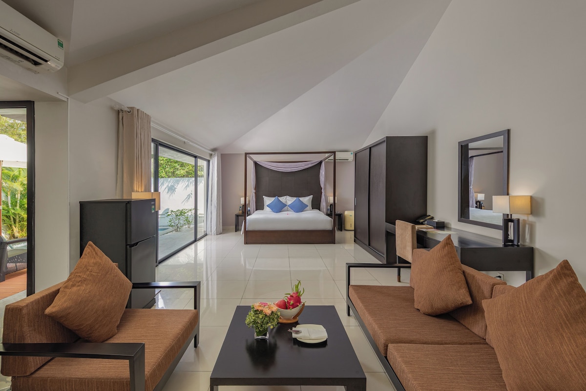 A modern hotel room with a large bed, two sofas, a coffee table with a fruit bowl, and a desk with a lamp.