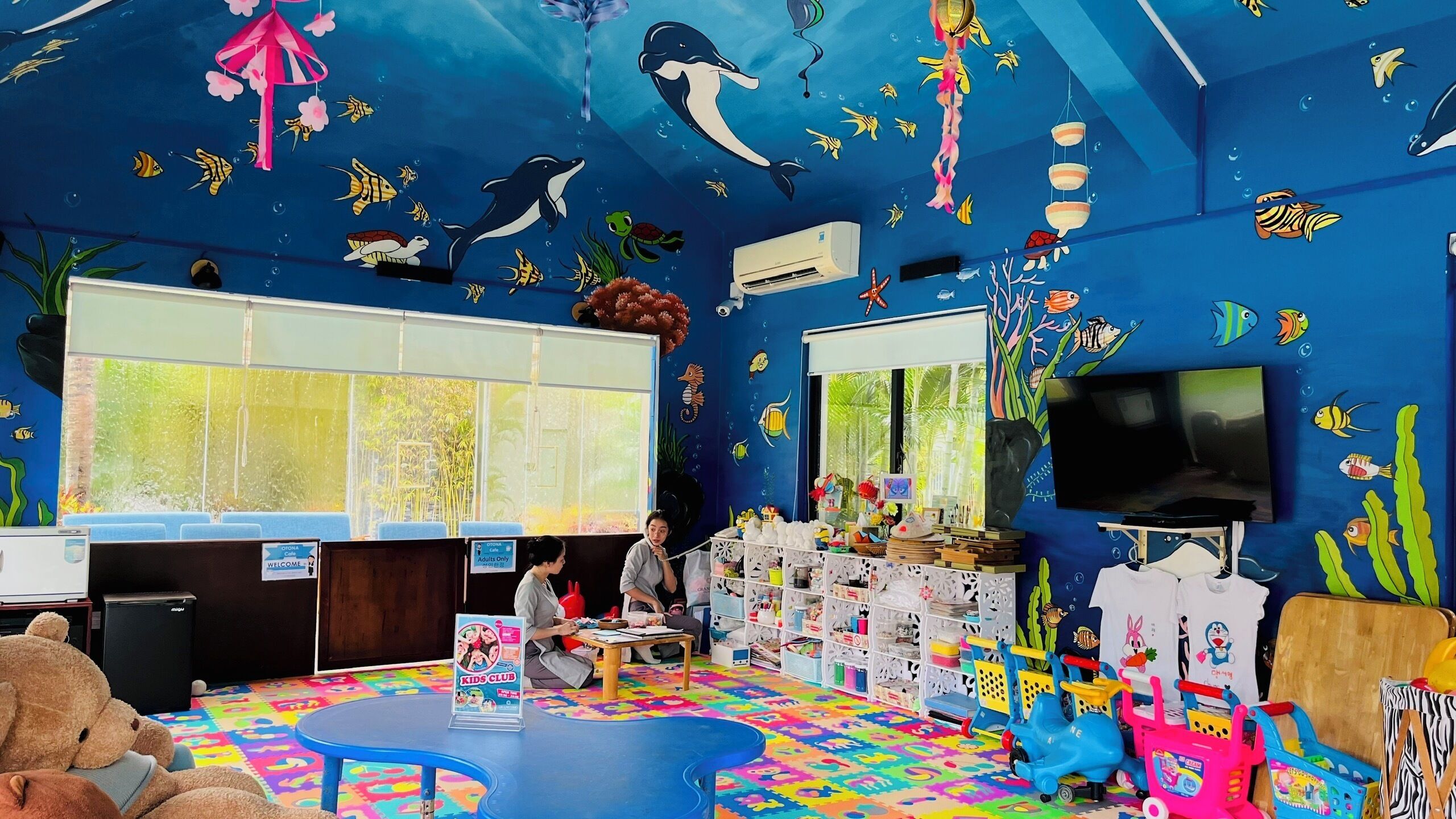 A children's playroom with a blue ocean-themed mural, a large window, and various toys and play equipment.