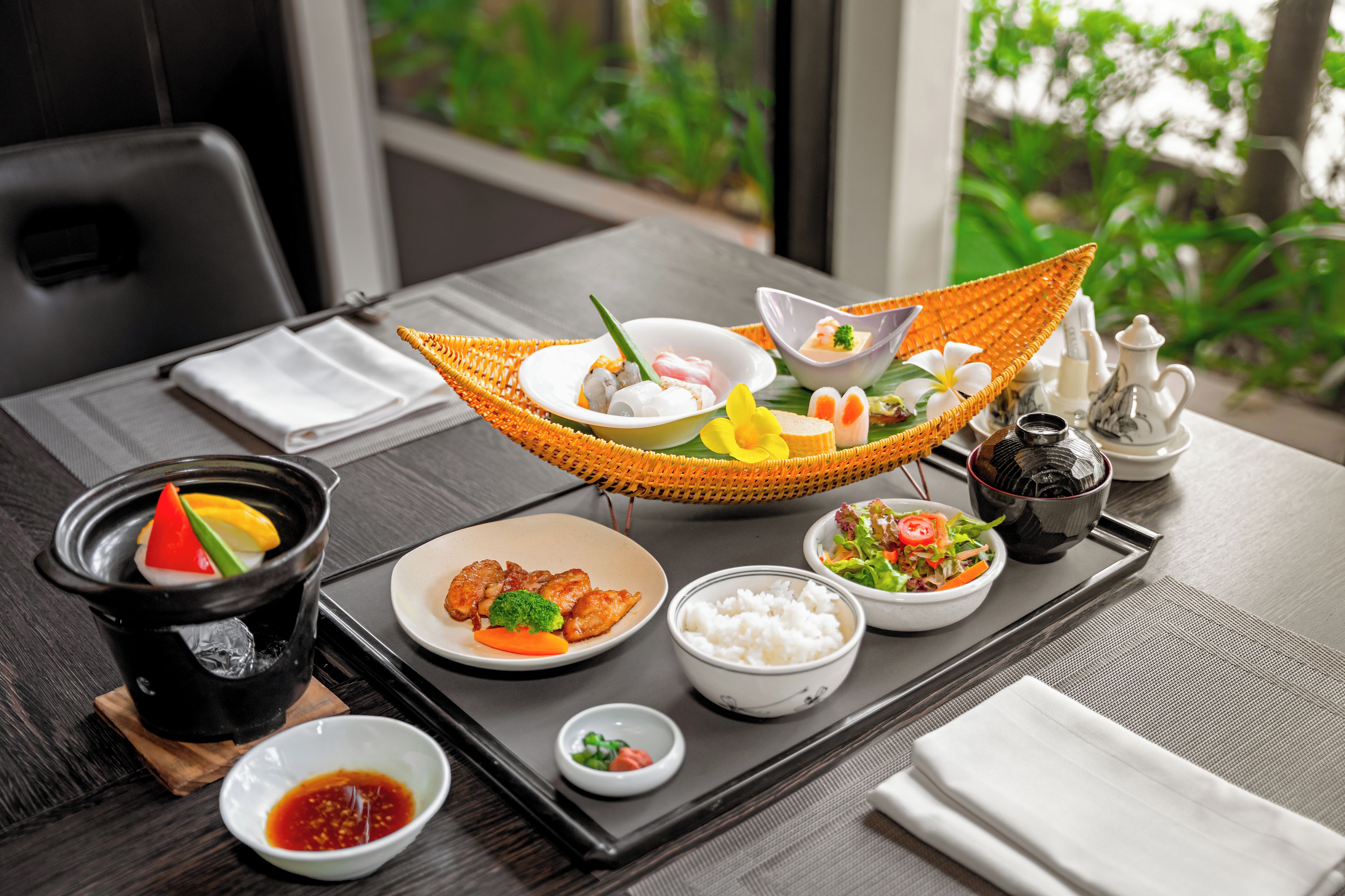 A dining table set with various dishes, including sushi, rice, and a salad, with a view of greenery outside.