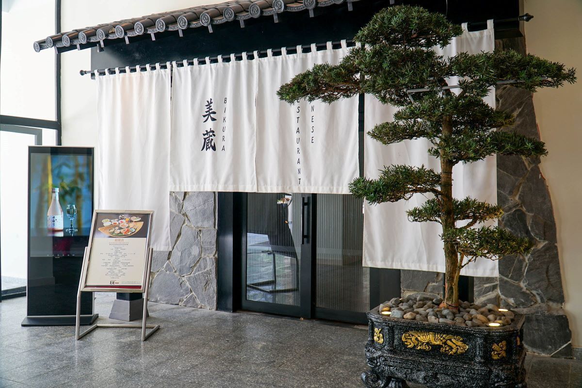 Entrada de un restaurante japonés tradicional con un letrero, un atril con la carta y un bonsái.