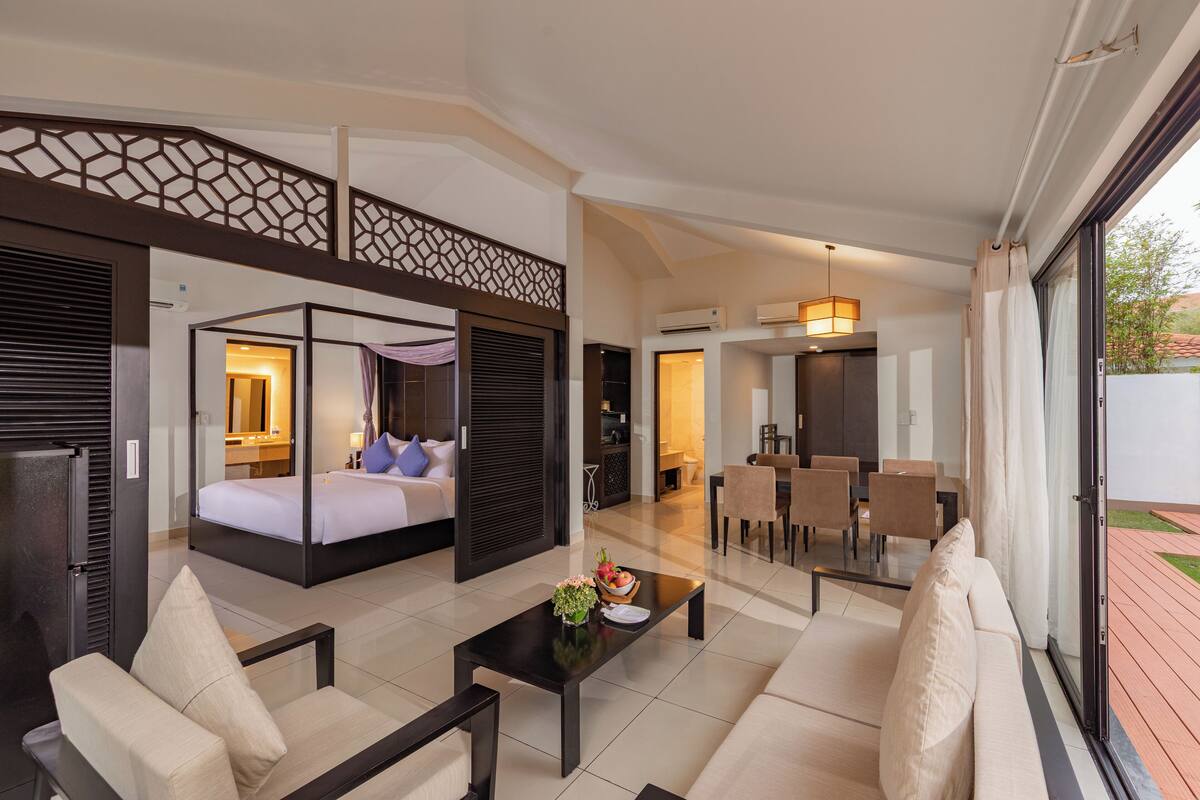 A modern hotel room with a large bed, a sofa, a coffee table, and a dining area with chairs.
