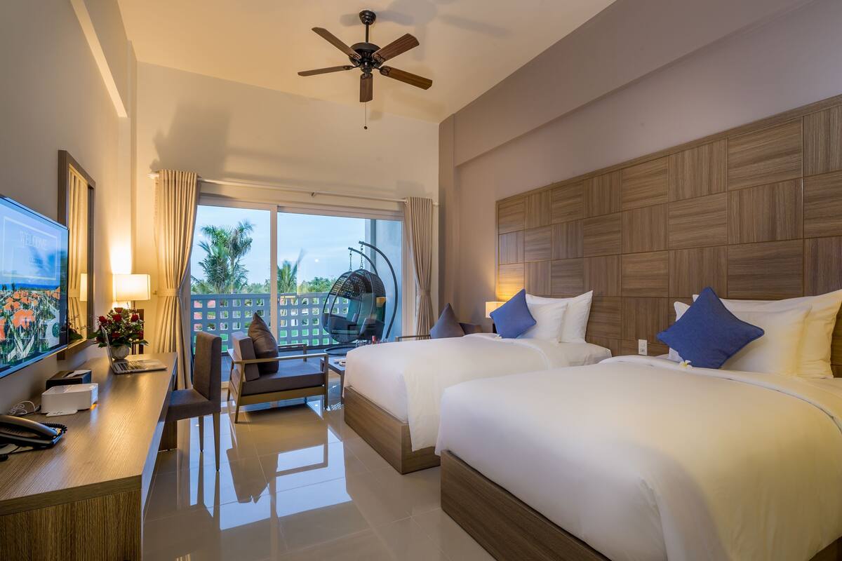 A hotel room with two beds, a desk with a laptop, a TV, and a balcony with a view of palm trees.