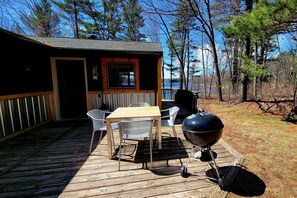 Eine Holzterrasse mit Tisch und Stühlen, einem Grill sowie einer Hütte mit Fenster und Tür.