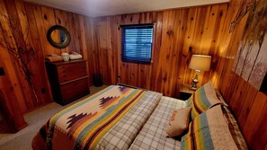 Ein Schlafzimmer mit Holzwänden, einem Bett mit farbenfroher Steppdecke, einem Schminktisch, einer Lampe und einem Fenster mit Jalousien.