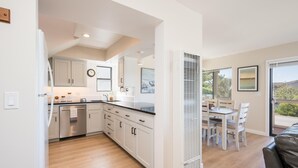 Een moderne keuken met witte kastjes, een zwart aanrecht en roestvrijstalen apparatuur. Er is een eethoek met een tafel en stoelen, en een groot raam dat uitzicht geeft op de omgeving.