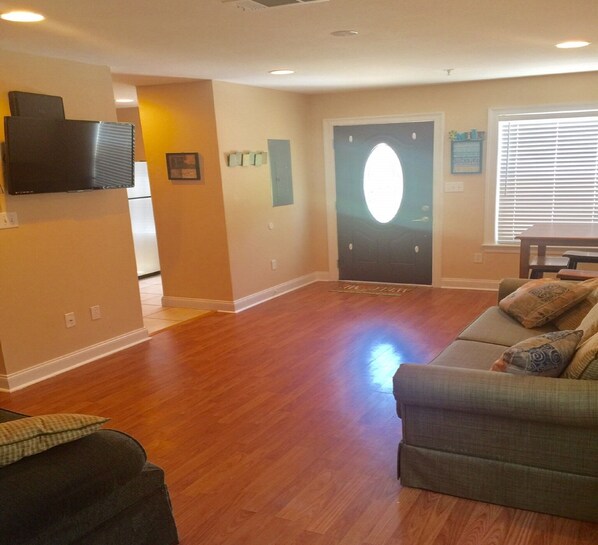 A spacious living room with hardwood flooring, a gray sofa with patterned cushions, a flat-screen TV mounted on the wall, and a door with a frosted glass window.