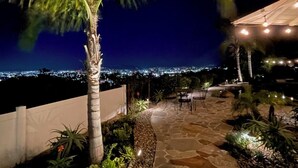 Ein gemütlicher Außenbereich mit Terrasse bei Nacht, einem Steinweg, Palmen und Lichterketten.