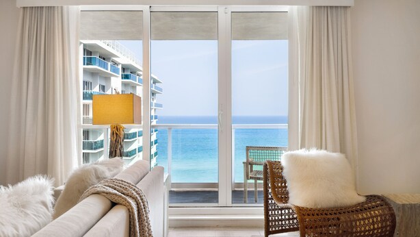 Habitación de hotel con balcón grande, sofá, sillón de mimbre y vista al océano y un edificio al otro lado del agua.