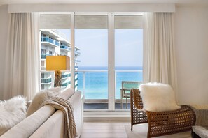 Habitación de hotel con balcón grande, sofá, sillón de mimbre y vista al océano y un edificio al otro lado del agua.