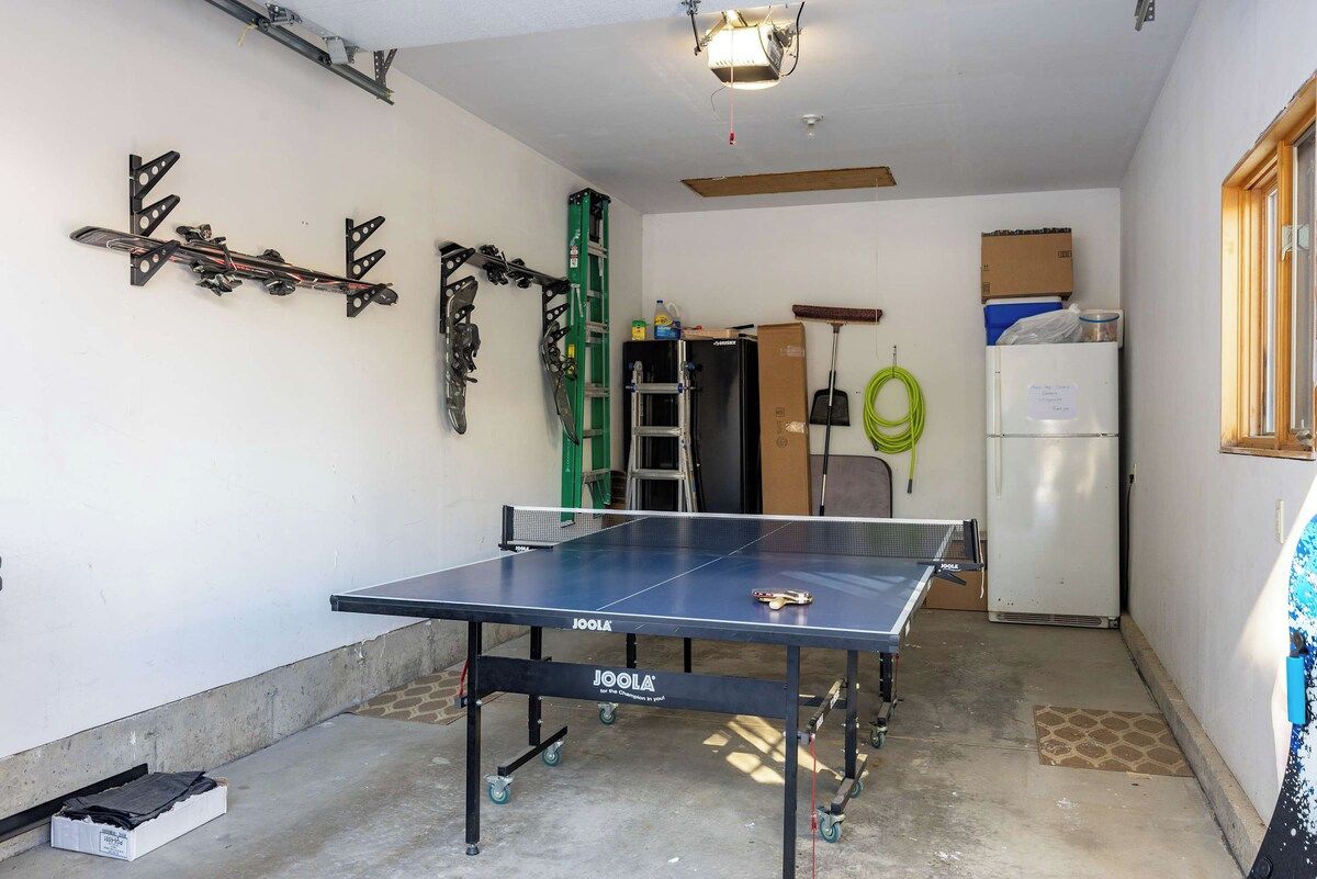 Una mesa de ping pong en una habitación con una escalera, un refrigerador y una puerta.