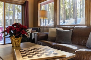 A cozy living room with a chessboard, a vase of red flowers, and a view of a forest through large windows.