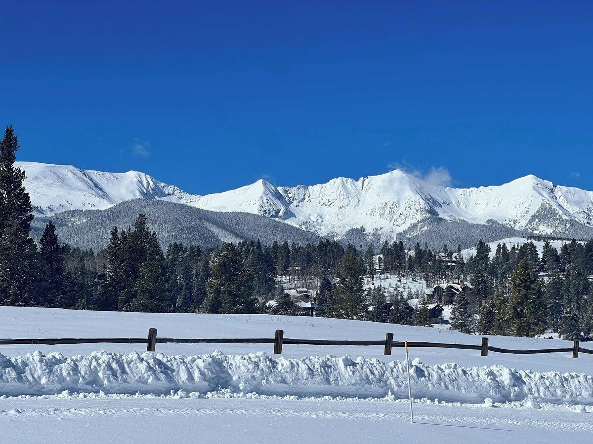 A snow-covered landscape with a wooden fence, mountains, and a clear sky.