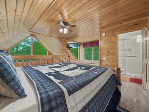 A bedroom with a wooden ceiling, a bed with a patterned bedspread, a ceiling fan, and a view of the outdoors.