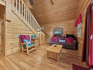 Interior de una cabaña de madera con una escalera, un sofá, un sillón mecedor y una mesita.