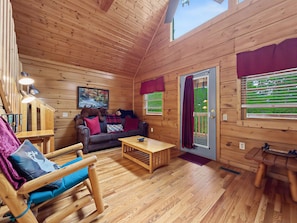 L’intérieur d’un chalet accueillant, avec un plancher en bois, un canapé avec des coussins rouges, un fauteuil à bascule, une petite table et une porte donnant sur l’extérieur.