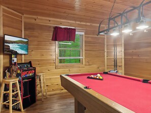 A pool table with a red felt surface, a pool cue, and a set of balls. There is a wooden stool and a video game machine with a cartoon character on the wall. A window with blinds and a view of greenery is visible.
