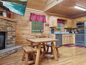 Interior de una cabaña de madera con chimenea de piedra, mesa de comedor de madera, zona de cocina y vista al océano a través de una ventana.