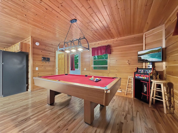 Une table de billard dans une pièce en bois, avec un ventilateur au plafond, un téléviseur et un petit coin bar.