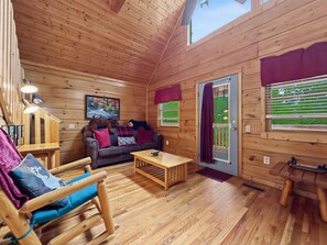 Un acogedor interior de cabaña con piso de madera, un sofá con cojines rojos, una mecedora, una mesita y una puerta que da al exterior.