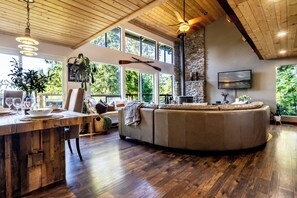 Un salón amplio con techo de madera, ventanas grandes, chimenea de piedra y televisor instalado en la pared.