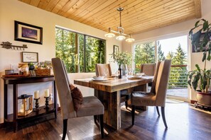 Un comedor con mesa de madera, sillas y ventanas amplias que dan a vista de árboles.
