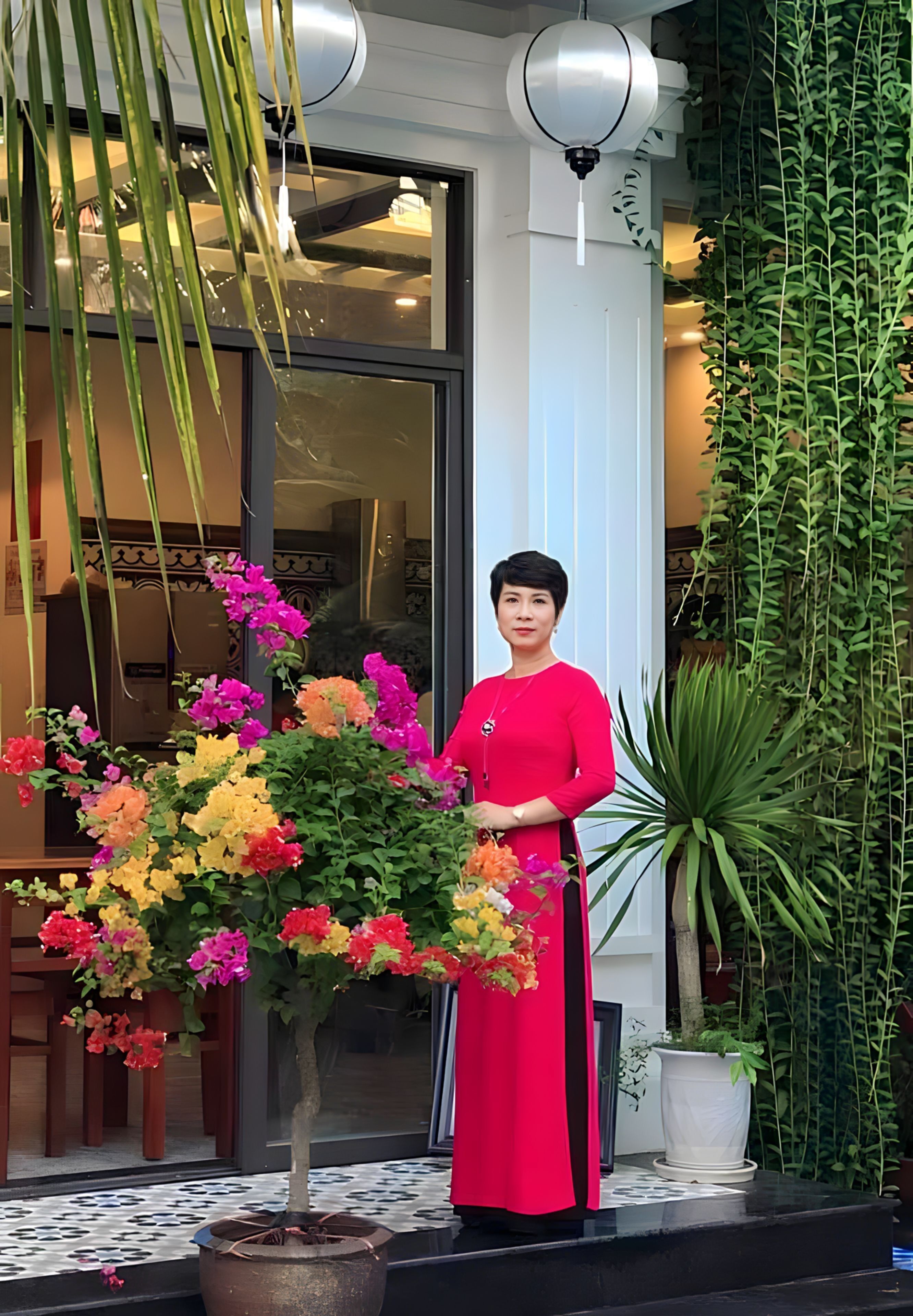 A woman in a red dress standing next to a vibrant flower arrangement in front of a building with large windows and indoor lighting.