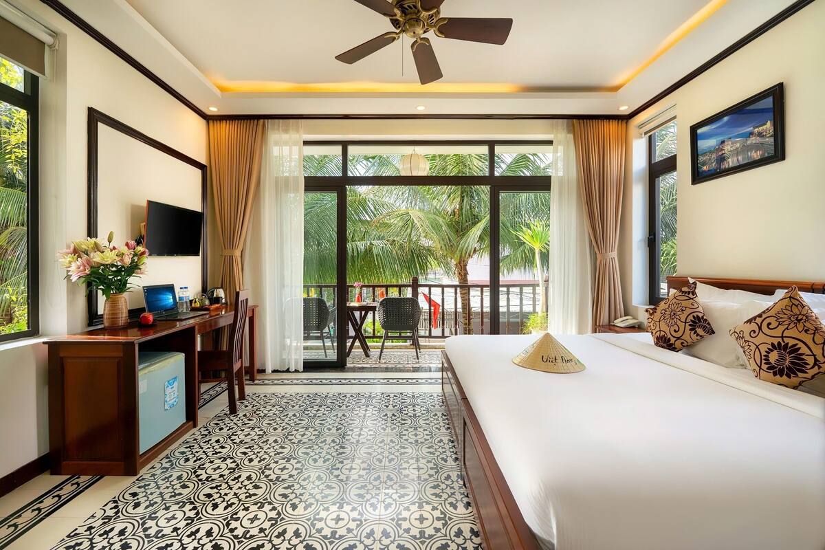 A hotel room with a bed, a desk with a laptop, a TV, and a balcony with a view of palm trees.
