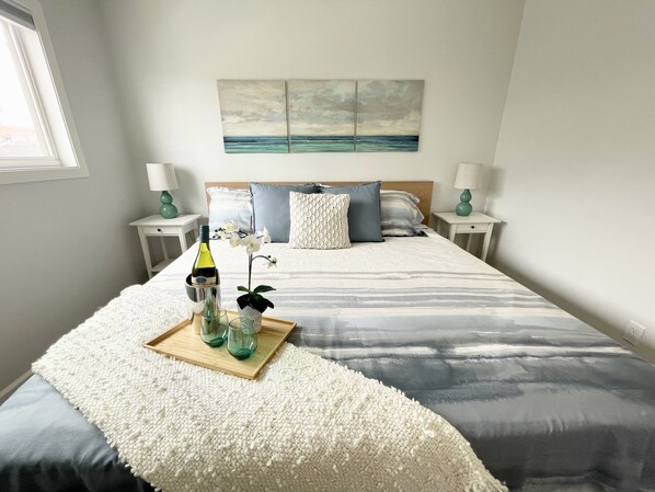 Un dormitorio ordenado con cama, mesita de noche con lámpara, una pequeña mesa con una botella y vasos, y un cuadro en la pared.