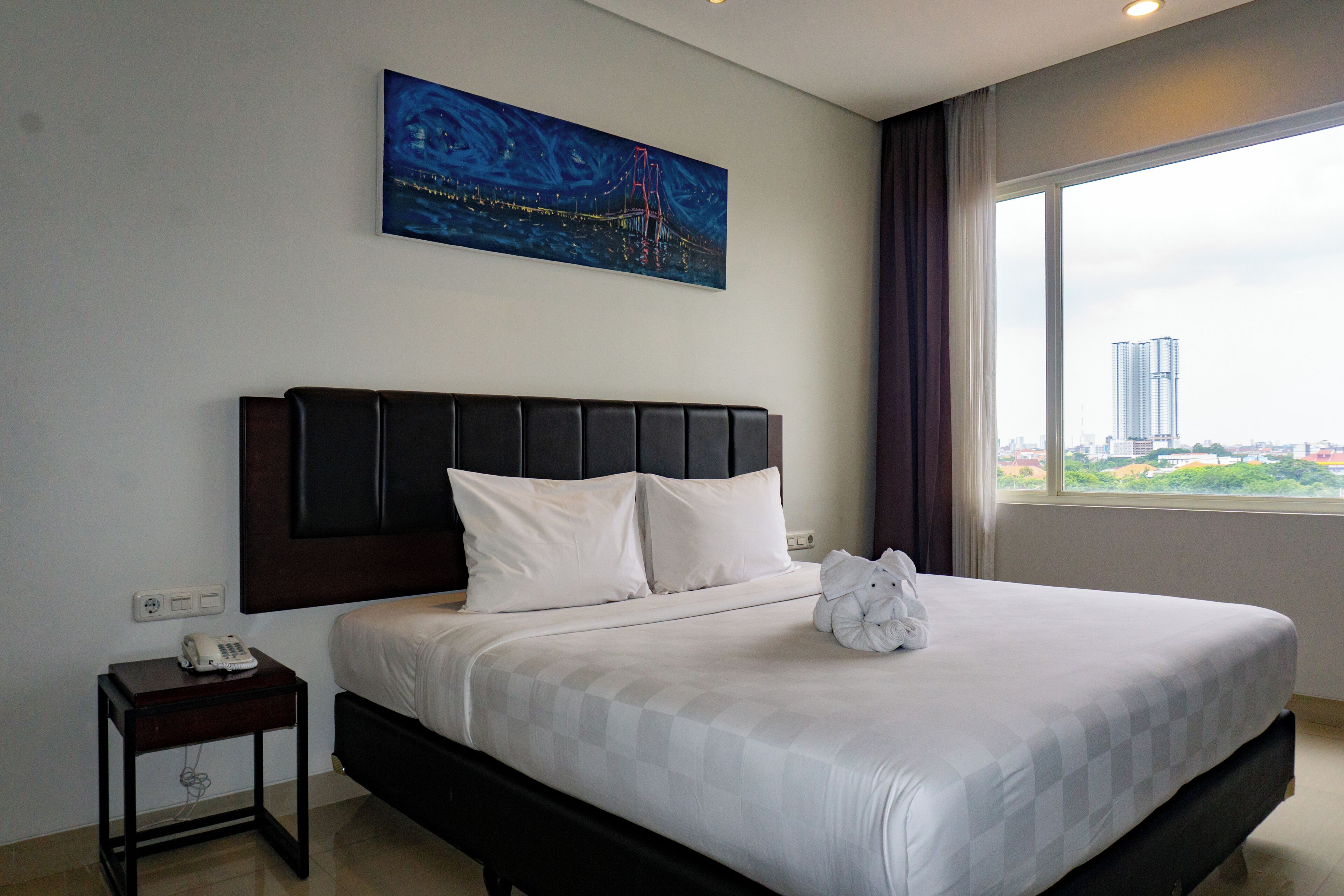Habitación de hotel con una cama, un cuadro, vista a una ciudad y un teléfono en la mesita de noche.