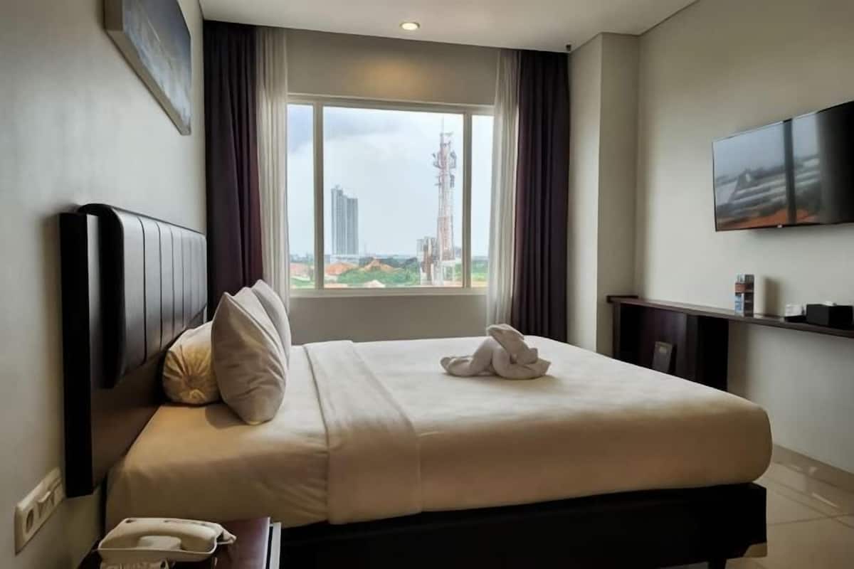 A hotel room with a bed, a bedside telephone, a window with curtains, and a flat-screen TV mounted on the wall.