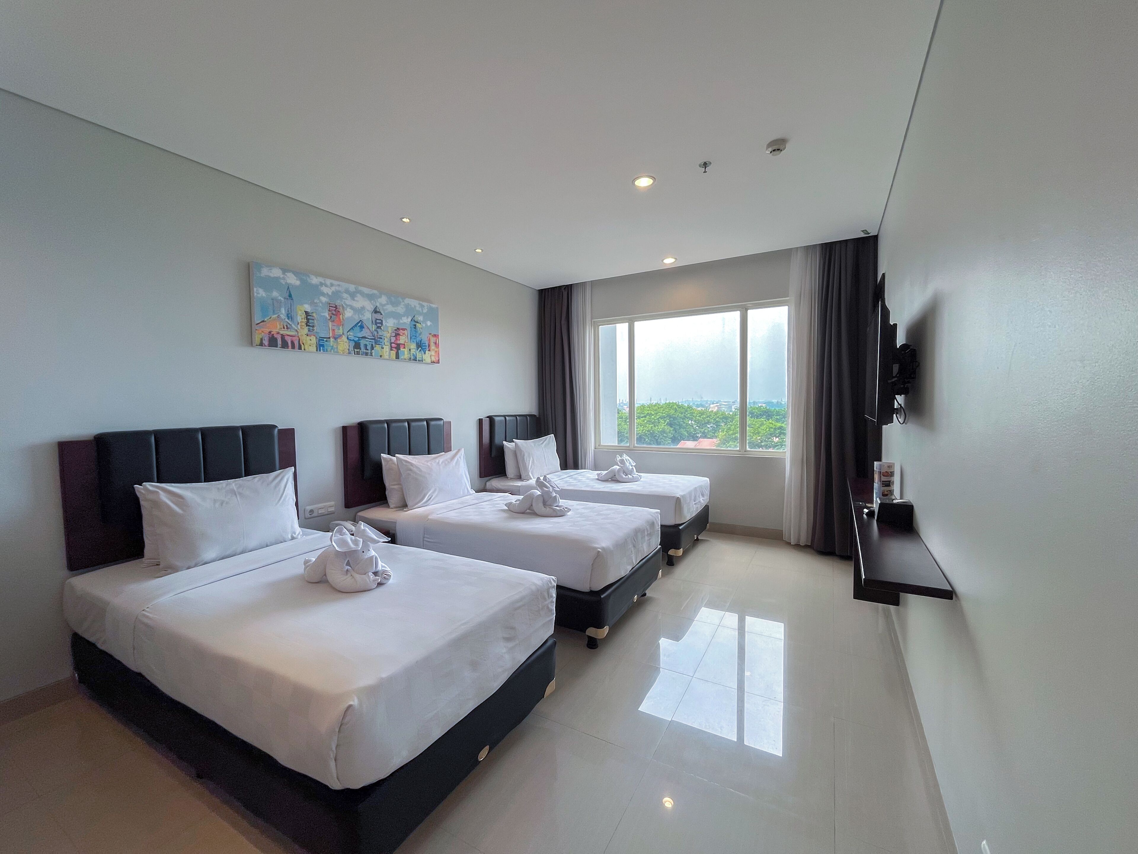 A hotel room with three beds, a painting on the wall, a TV mounted on the wall, and a large window with a view.