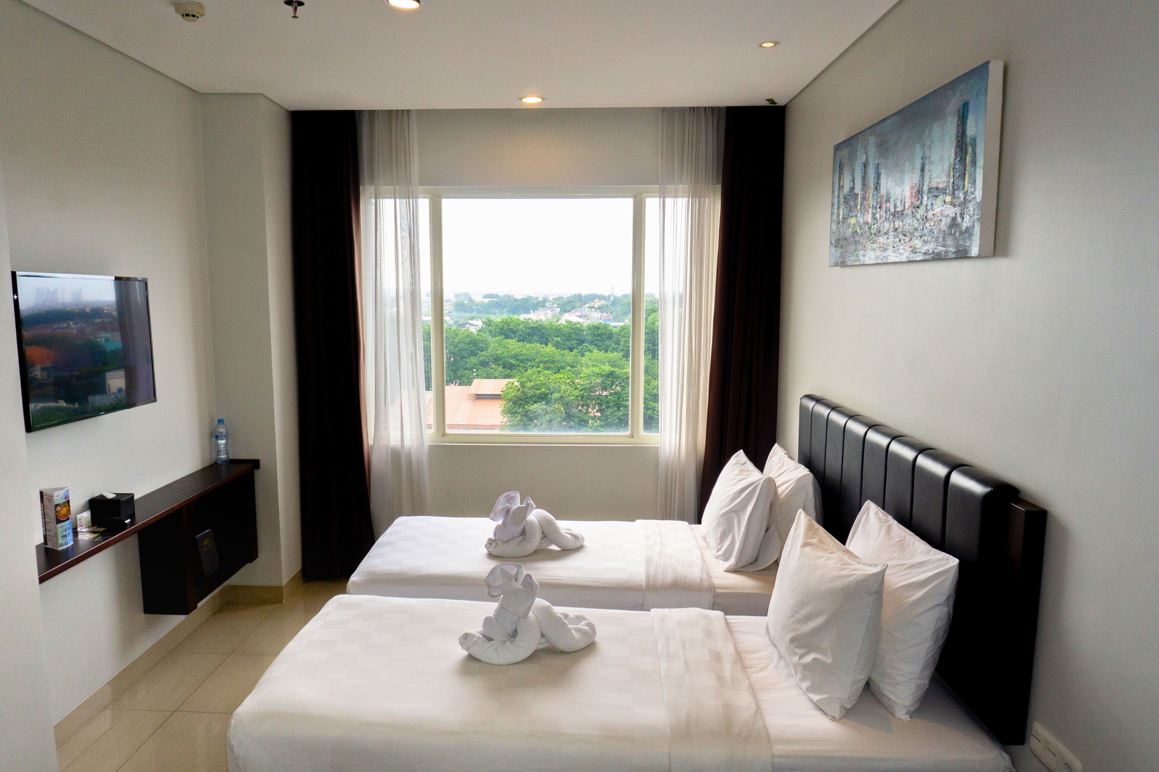 A hotel room with two beds, a flat-screen TV, a painting, and a window with a city view.