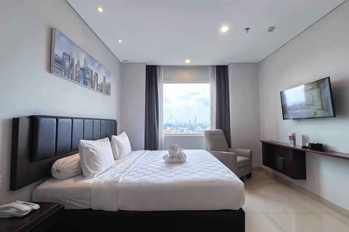 A hotel room with a bed, a TV, a chair, a desk, and a window with a city view.