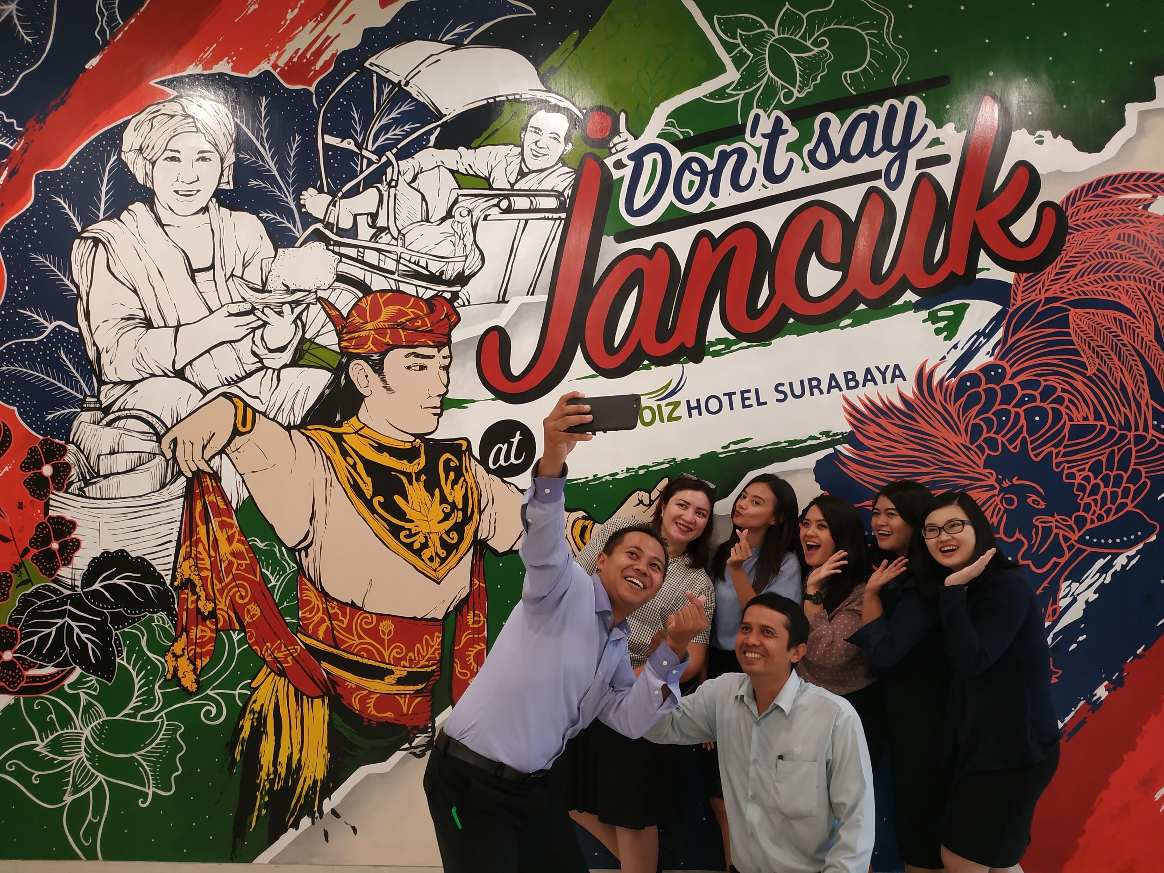 Un groupe de personnes posant devant une fresque murale sur laquelle figure une pancarte bien visible avec l’inscription « Ne dites pas Jancuk » et le nom d’un hôtel.
