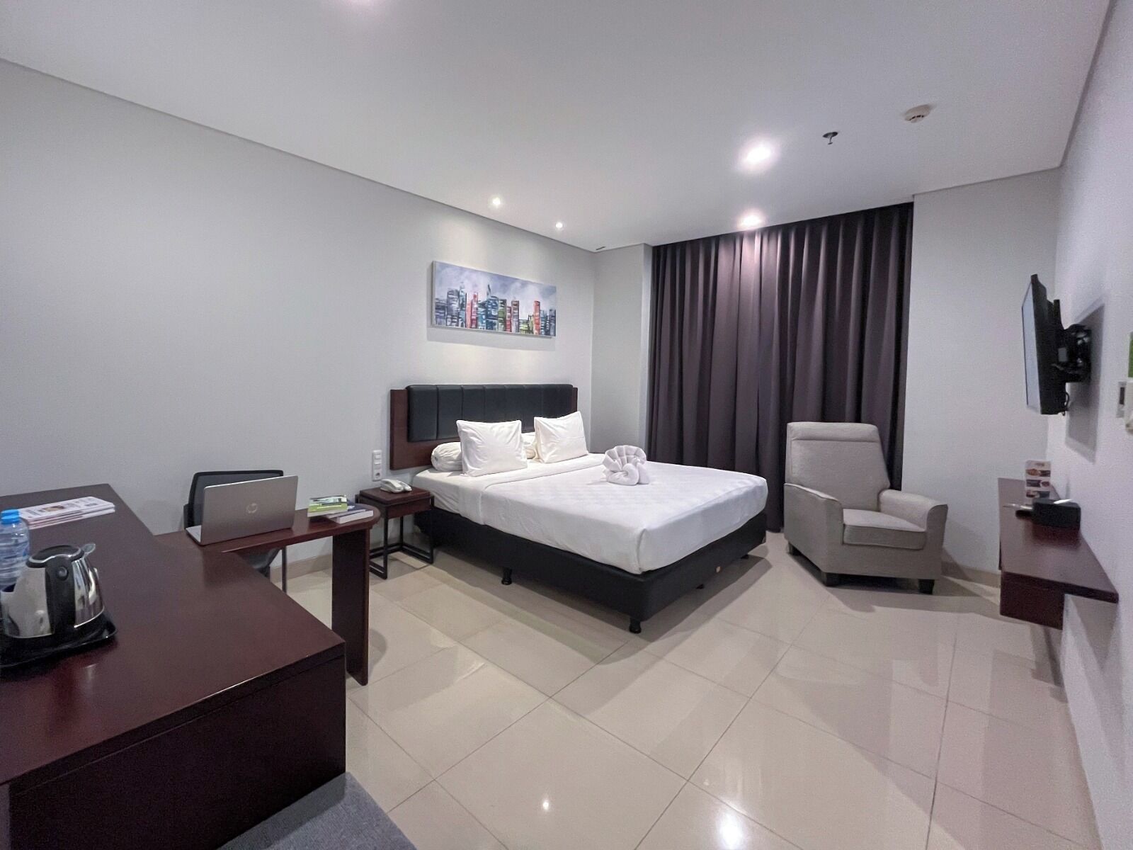 A hotel room with a bed, desk, chair, and a cityscape painting on the wall.