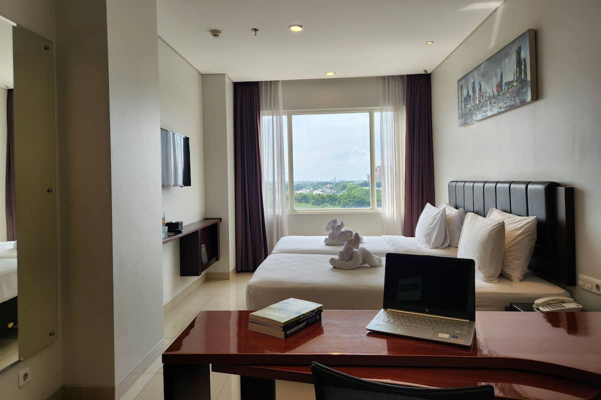 A hotel room with a bed, a desk with a laptop, a bedside table with books, a telephone, and a painting on the wall.