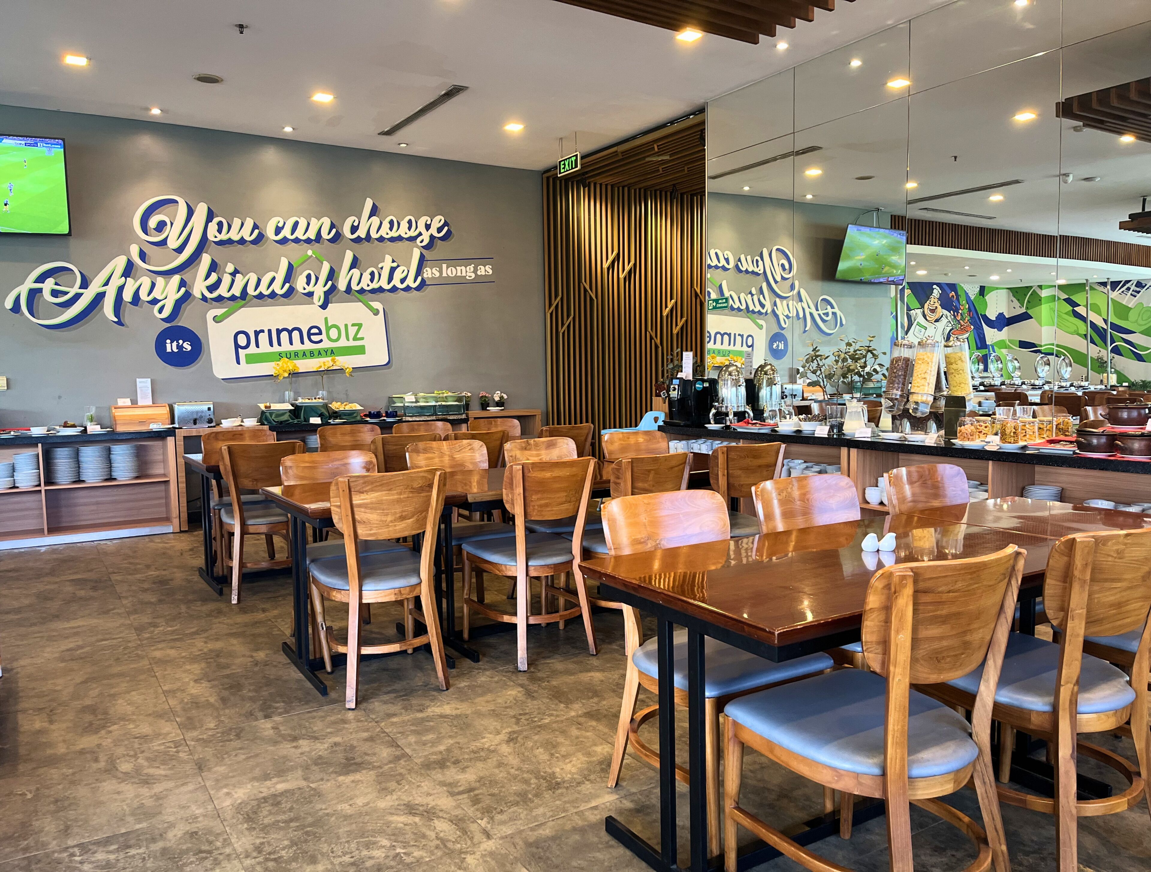 A hotel dining area with wooden chairs and tables, a buffet counter with food items, and a wall with a sign that reads 'You can choose any kind of hotel as long as primebiz.'