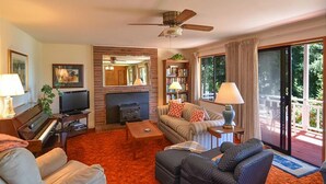 A cozy living room with a fireplace, a television, a piano, and a balcony with a view of greenery. - San Juan Island, Roche Harbor Heights #122 (Friday Harbor)